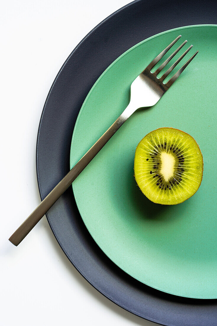 From above of half of fresh ripe juicy kiwi and fork placed on grey and green plates on white surface