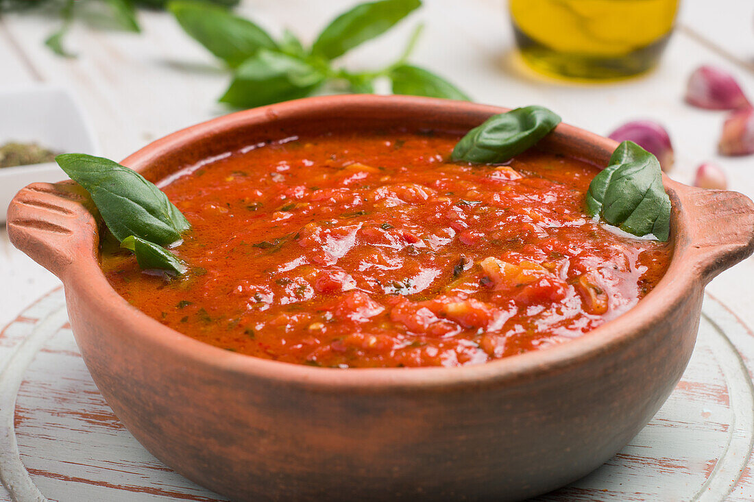 Holzschüssel mit roter Marinara-Soße aus Tomaten und Basilikumblättern auf dem Tisch, dazu Olivenöl und Knoblauch