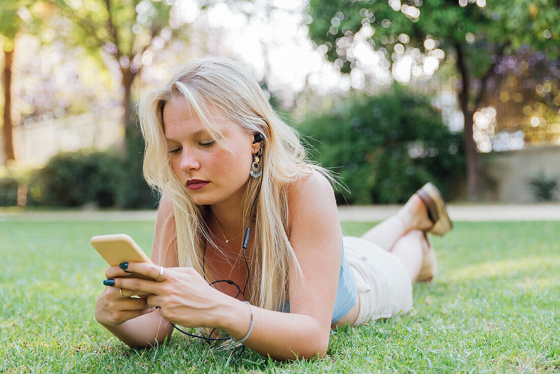 Unaufgeregte, charmante Frau, die im Sommer im Gras im Park liegt und mit Kopfhörern Musik hört