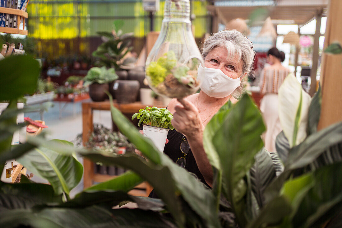 Weibliche Einkäuferin mittleren Alters mit einer Gesichtsmaske aus Stoff schaut weg, während sie auf eine Pflanze in einem Florarium in einem Gartencenter zeigt