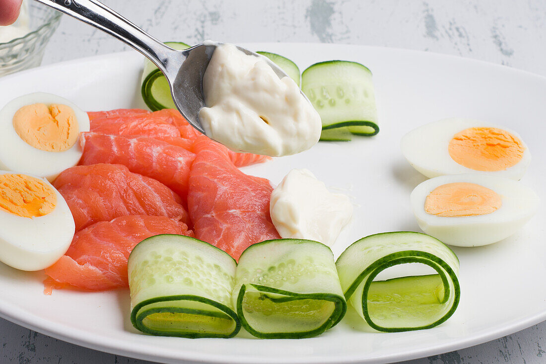 Unbekannter Koch gibt Frischkäse auf einen Teller mit Lachs, Eiern und Gurken auf dem Tisch