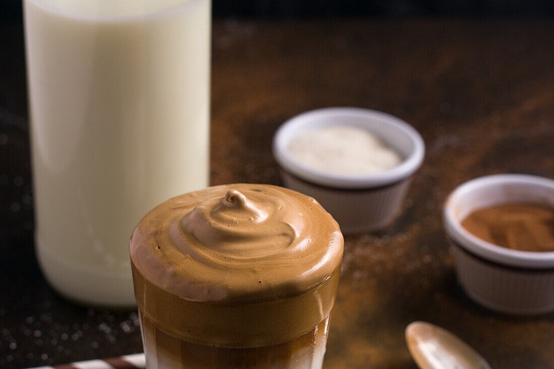 Ein Glas köstlicher Dalgona-Kaffee mit Milch und schaumigem Topping steht auf einem schwarzen, unordentlichen Tisch mit Kakaopulver und Zucker