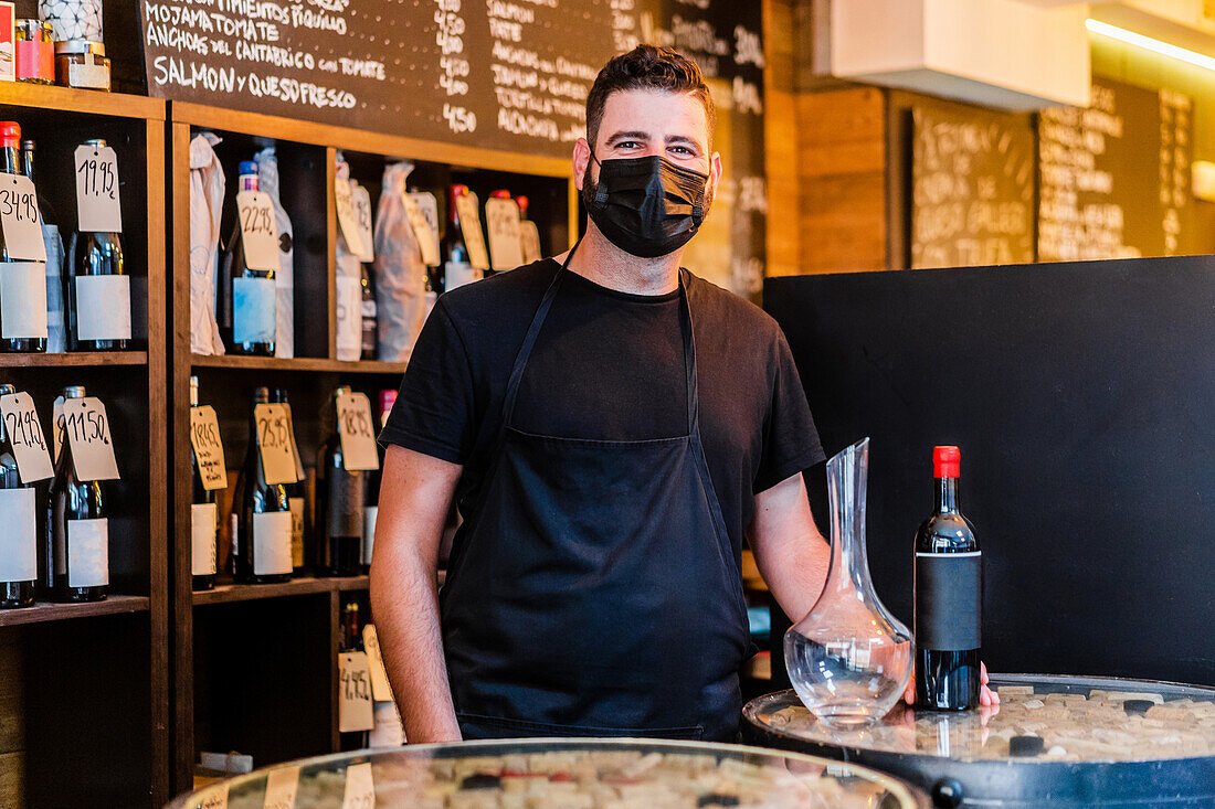 Bärtiger ethnischer Barmann schaut in die Kamera neben einer Glasflasche mit Wein in der Nähe einer Vase auf einem Metalltablett auf dem Tresen