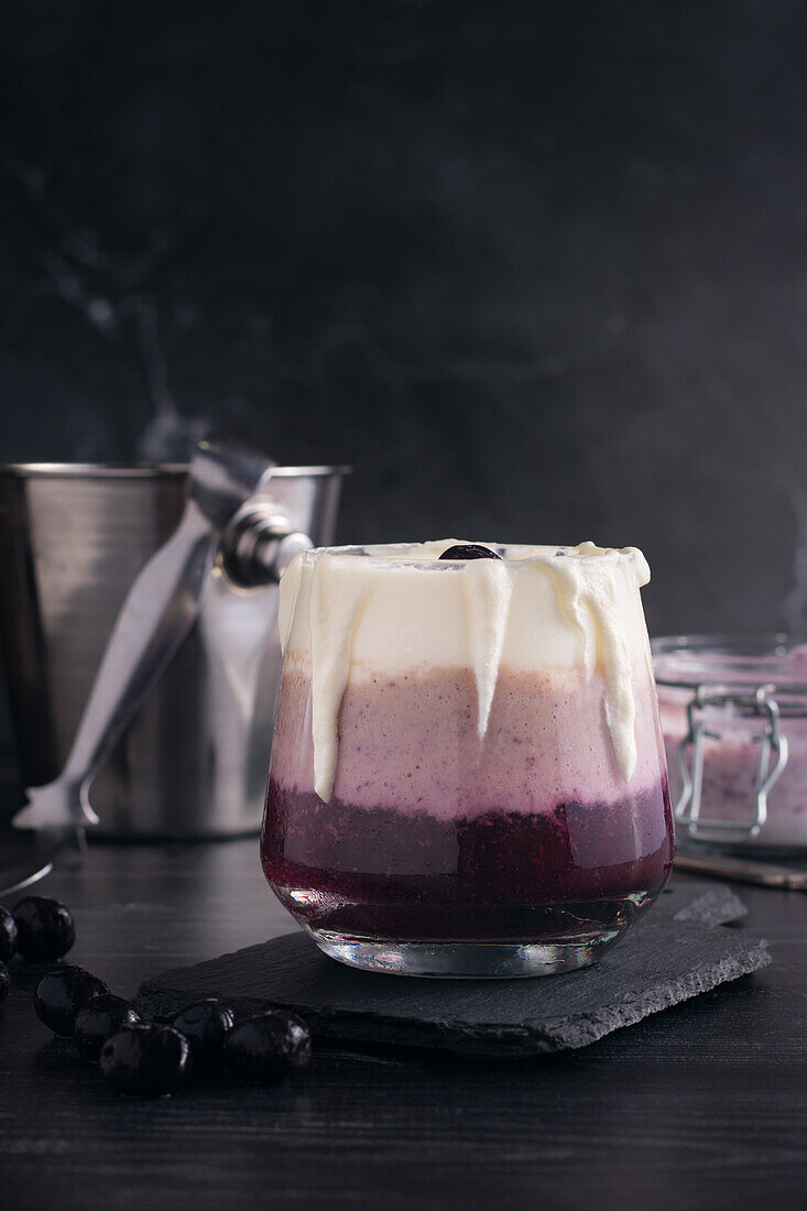 Glass of delicious banana and blueberry smoothie with whipped cream on chopping board against jar of yogurt and metal bucket