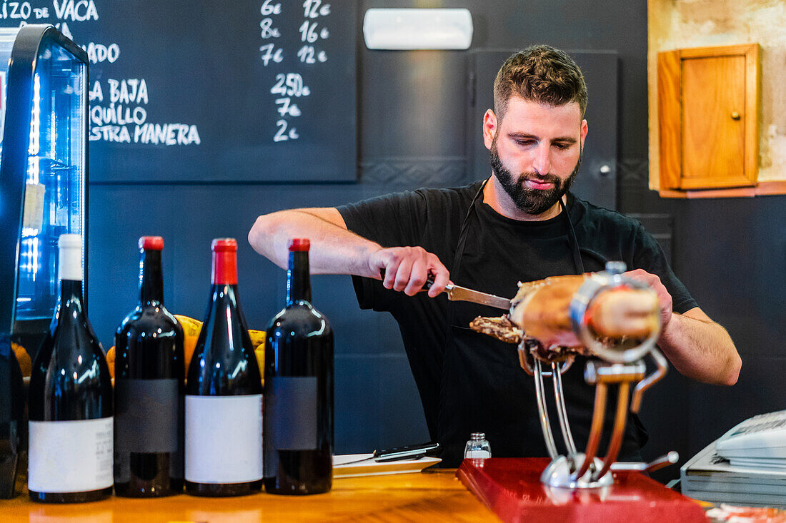 Bärtiger professioneller ethnischer Schneider bereitet leckeren Jamon in einem Restaurant mit Weinflaschen zu