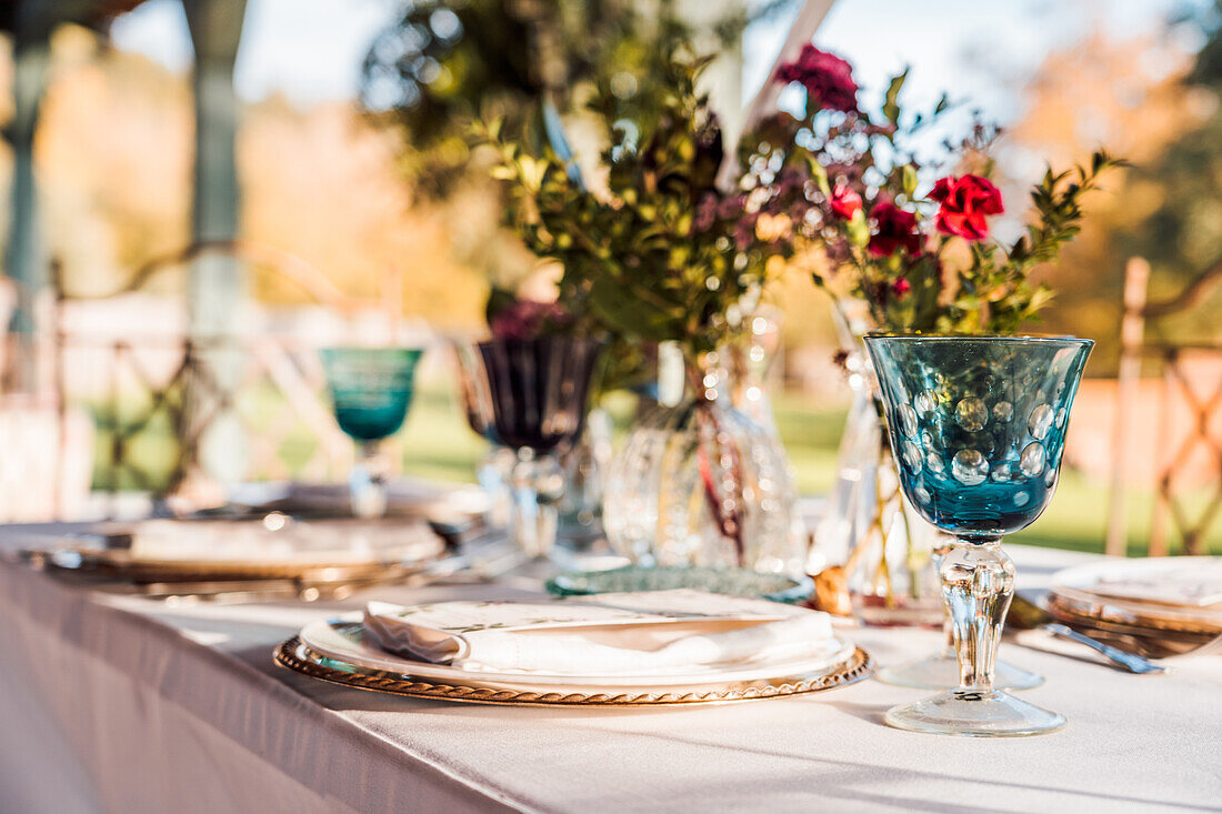 Nahaufnahme einer gedeckten Festtafel mit Kristallgläsern, Besteck und Serviette auf einem Teller neben einem Strauß frischer Blumen für eine Hochzeit und einer Menükarte