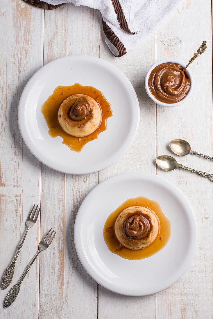 Top view of egg custards topped with sweet Dulce de leche served on white plates on table with cutlery in kitchen