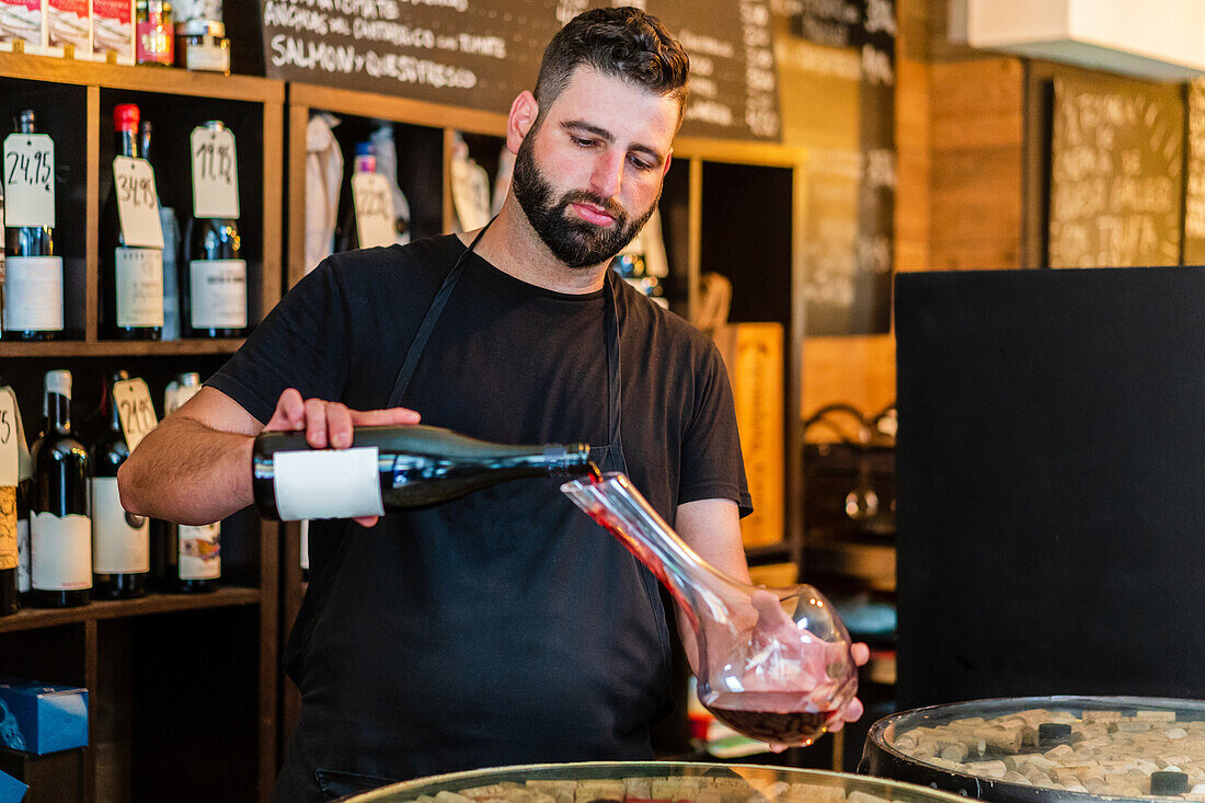 Fokussierter männlicher Sommelier mit Bart und Schnurrbart in schwarzer Schürze steht am Bartresen und gießt eine Flasche Rotwein in eine Glaskaraffe in einem Restaurant