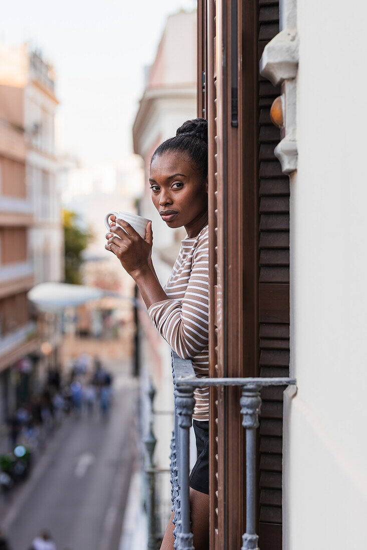 Side view of reflective African American female with cup of hot beverage looking at camera from balcony in city