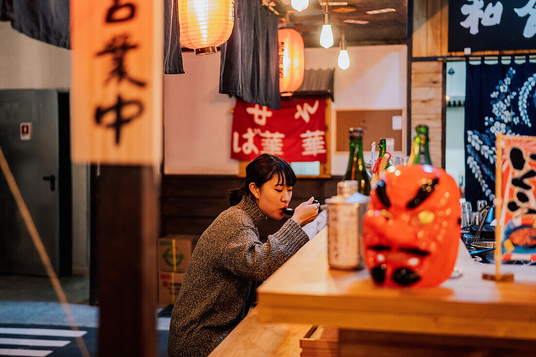 Young Asian woman in sweater eating ramen with spoon at wooden counter in cafe