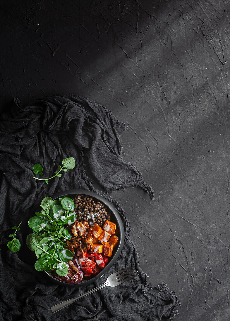 Draufsicht auf Salat mit Kürbis und Paprika, dekoriert mit grünen Basilikumblättern auf schwarzem Hintergrund