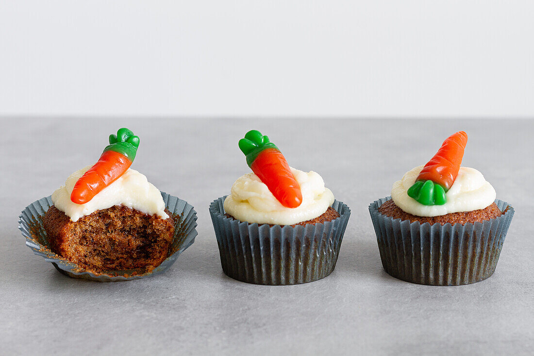 Reihen von süßen, leckeren Karotten-Cupcakes mit zarter Sahne und karottenförmigen Gummibärchen auf grauer Oberfläche