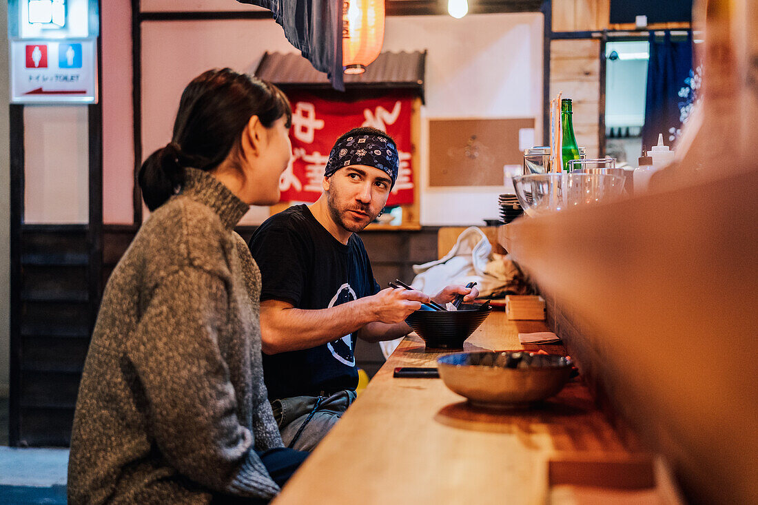 Seitenansicht einer Frau und eines Mannes, die sich beim Essen asiatischer Speisen an einem Holztresen in einem modernen Café unterhalten