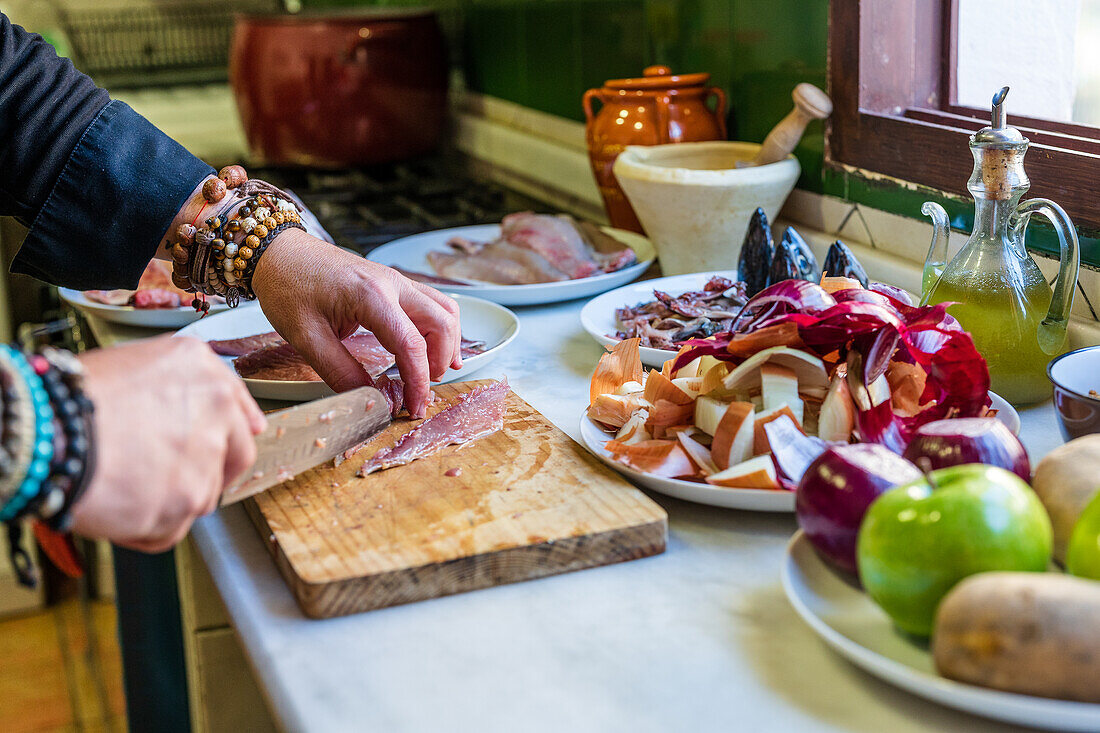 Abgeschnittener, nicht erkennbarer männlicher Koch in Uniform schneidet Fischfilet auf einem Brett, während er an einem Tisch mit verschiedenen Zutaten in der Küche steht