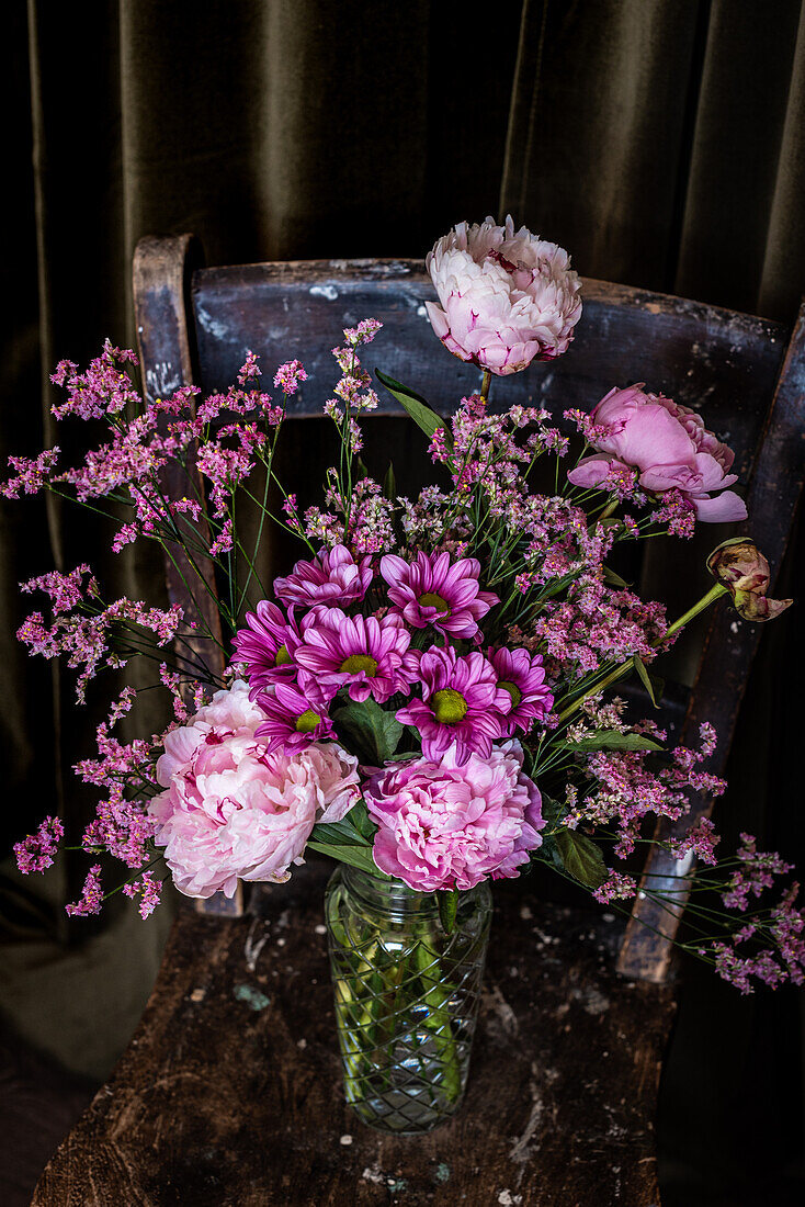Bouquet of fresh colorful peonies and chrysanthemums in glass vase placed on weathered wooden chair near curtains in light room