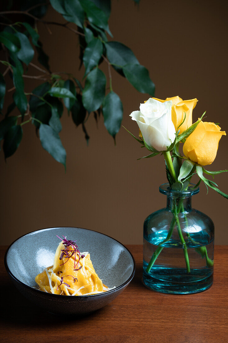Tortellini mit Käse und Walnüssen in einer schwarzen Schale, garniert mit Mikrogrün, neben einer Vase mit gelben Rosen.