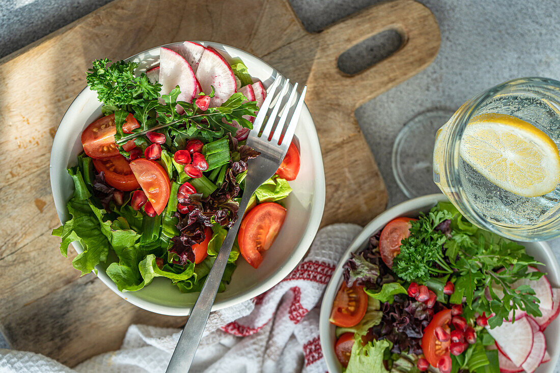 Nahaufnahme eines gesunden Gemüsesalats mit gemischtem Grün, Radieschen, Kirschtomaten und Granatapfelkernen, neben einem Glas Zitronenwasser auf einem Holzbrett