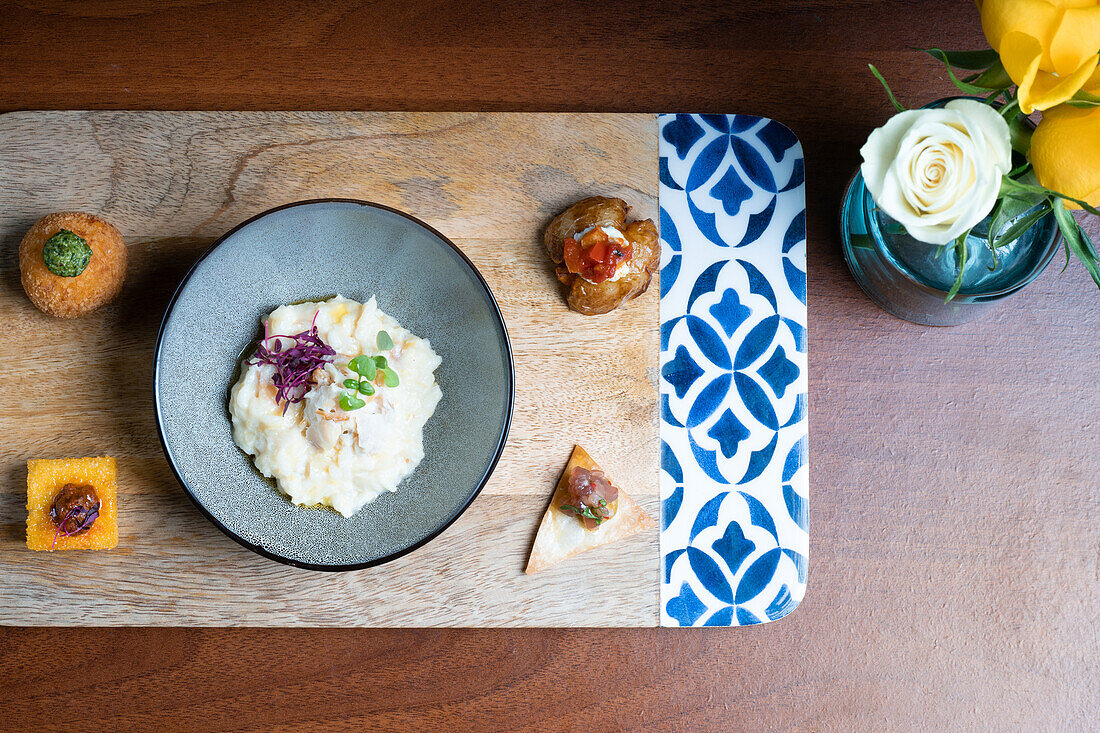 Von der obigen Auswahl an Gourmet-Häppchen, die eine Schale mit cremigem Risotto umgeben, das mit Mikrogrün verziert ist, präsentiert auf einem Holztisch mit Blumenakzent.