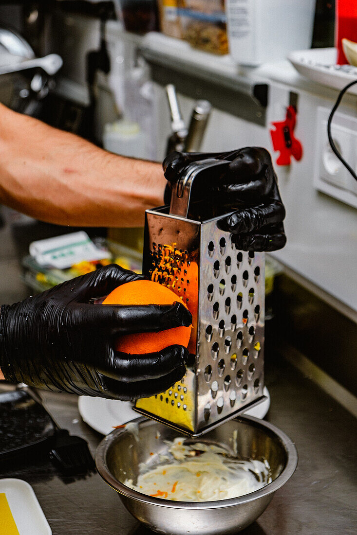 A chef wearing black gloves grates an orange over a metal bowl, preparing zest for a culinary recipe in a professional kitchen