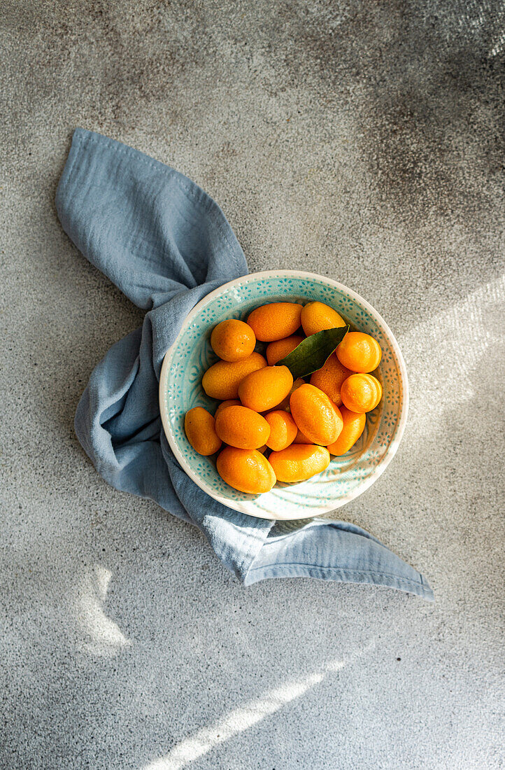 Frische Kumquats in einer dekorativen Schale mit einer blauen Serviette auf einem strukturierten Betonhintergrund (von oben)