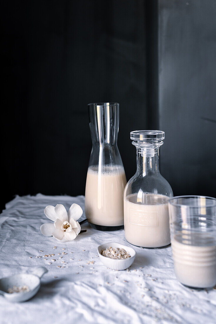 Two glass bottles of homemade oat milk with a glass half full and scattered oats on cloth