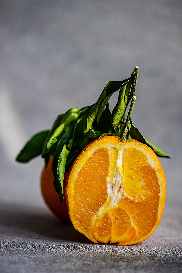 Eine leuchtende, frisch geschnittene Orange mit üppigen grünen Blättern vor einem strukturierten grauen Hintergrund, der die Saftigkeit und natürliche Schönheit der Frucht hervorhebt