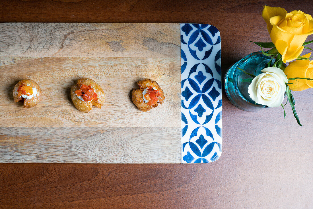 Von oben elegante Baby-Kartoffel-Häppchen mit geschmolzenem Mozzarella und rotem Pfeffer auf einer Holzplatte mit dekorativen Blumen.