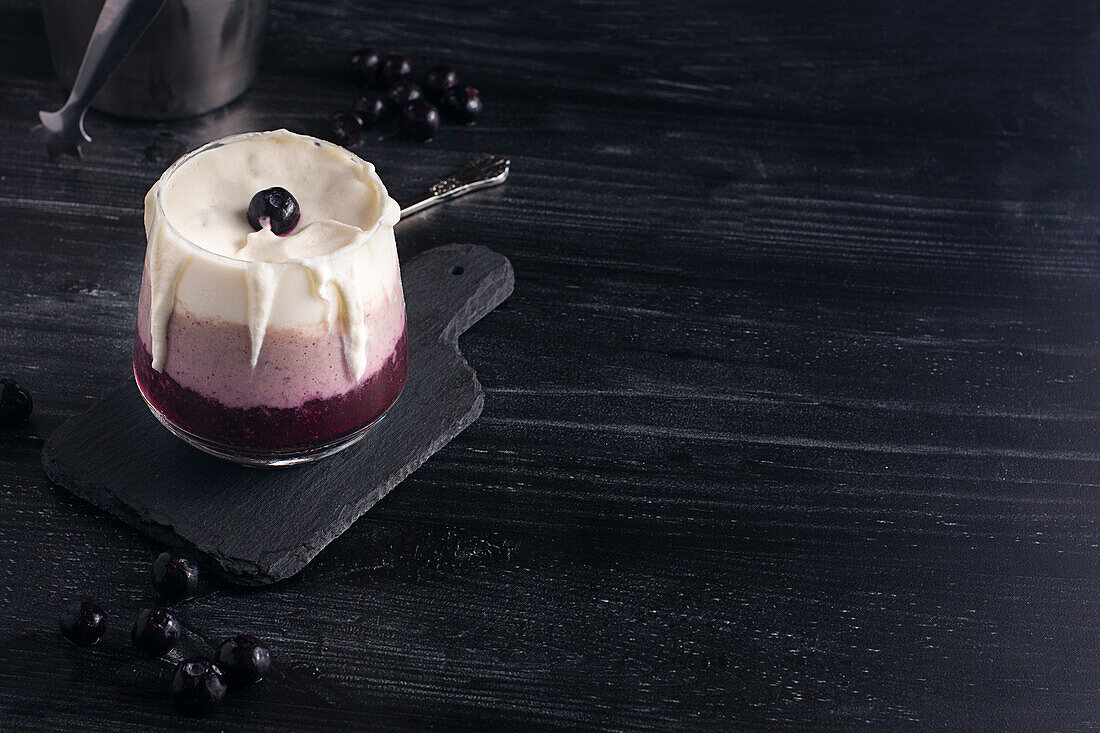 Glas köstlicher Bananen- und Blaubeer-Smoothie mit Schlagsahne auf Schneidebrett vor Joghurtglas und Metalleimer