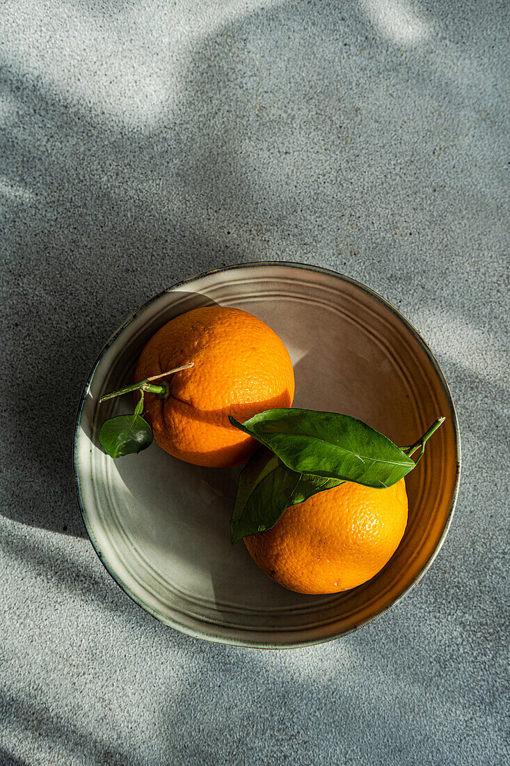 Ein Paar reife Orangen mit üppigen grünen Blättern sitzen in einer einfachen Schale und werfen Schatten auf einen strukturierten grauen Hintergrund