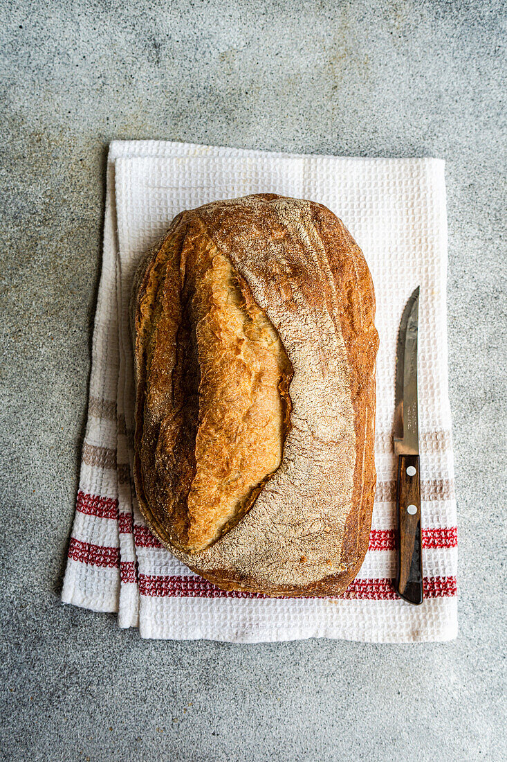 Draufsicht auf goldbraunes Roggensauerteigbrot auf einem weißen Tuch mit roten Streifen, daneben ein Brotmesser auf einer strukturierten Oberfläche