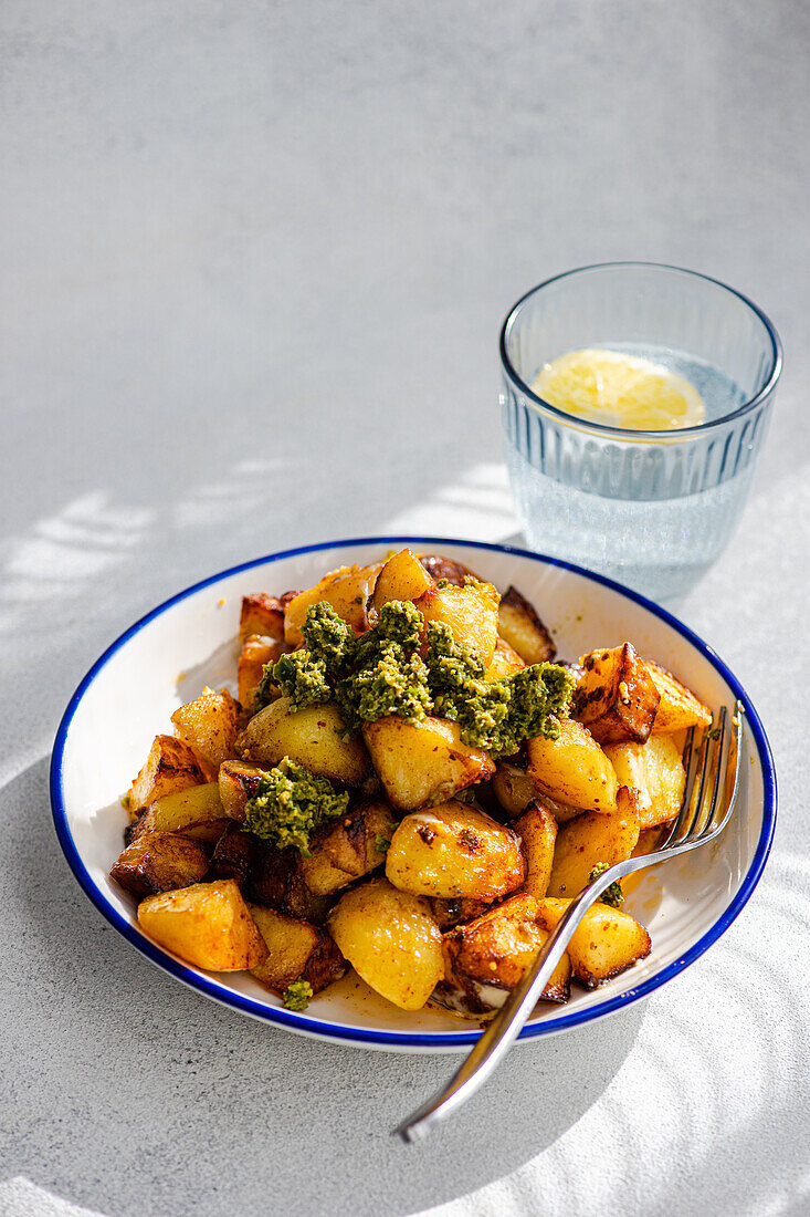 Roasted golden potatoes topped with vibrant green pesto on a white enamel plate with a blue rim, accompanied by a clear glass of water, all set upon a light grey textured surface