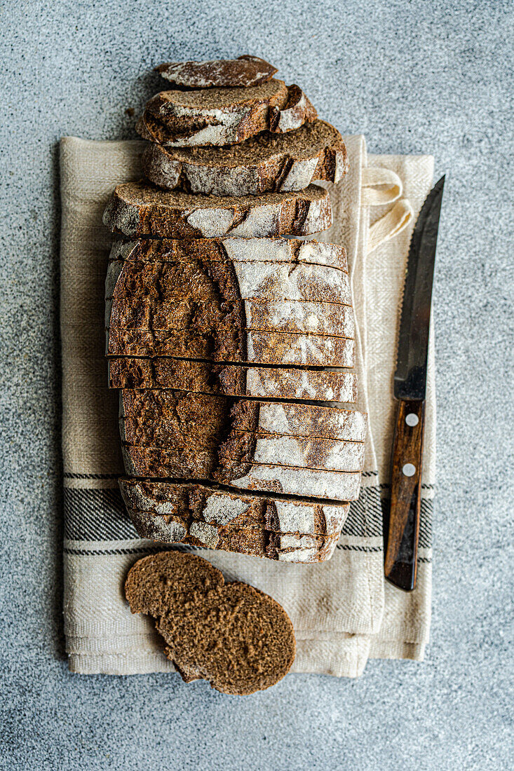 Blick von oben auf einen Laib Roggensauerteigbrot, teilweise aufgeschnitten, mit einem Brotmesser auf einem beigen Küchentuch vor einem strukturierten Hintergrund