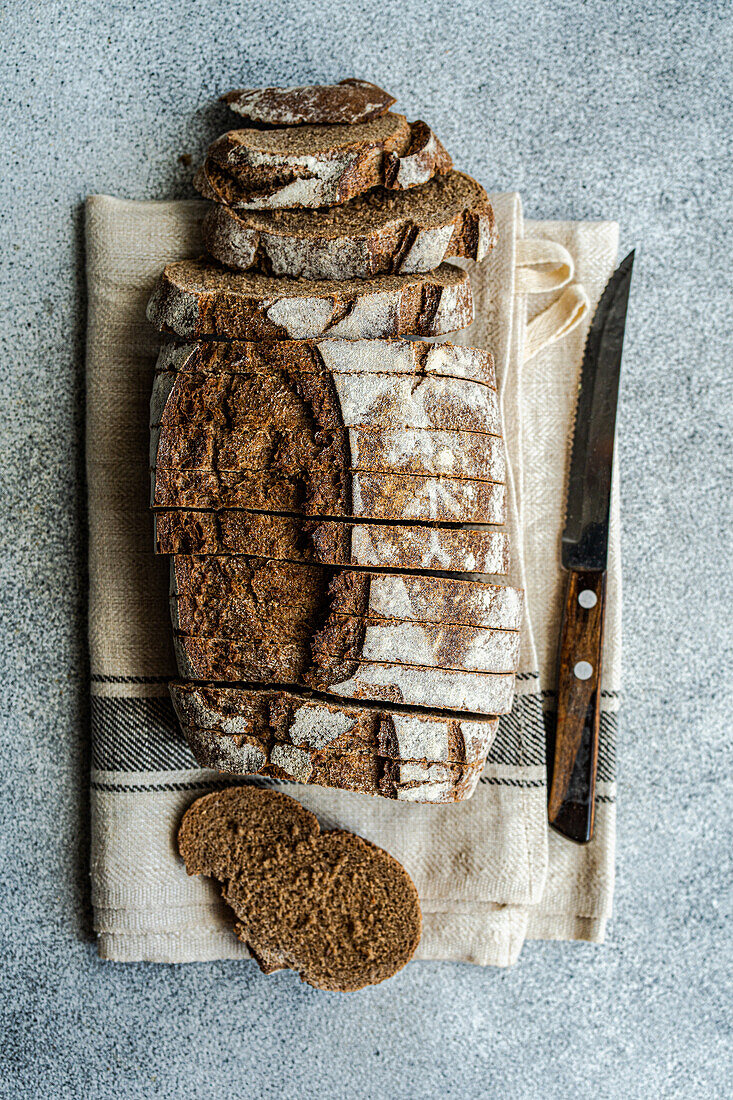 Top view of loaf of rye sourdough bread, partially sliced, with a bread knife on a beige kitchen towel against a textured backdrop