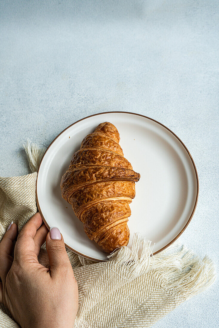 Draufsicht auf eine anonyme Person, die einen Teller mit einem frisch gebackenen Croissant auf einem strukturierten Tuch in der Hand hält und damit einen einfachen und gemütlichen Frühstücksmoment festhält