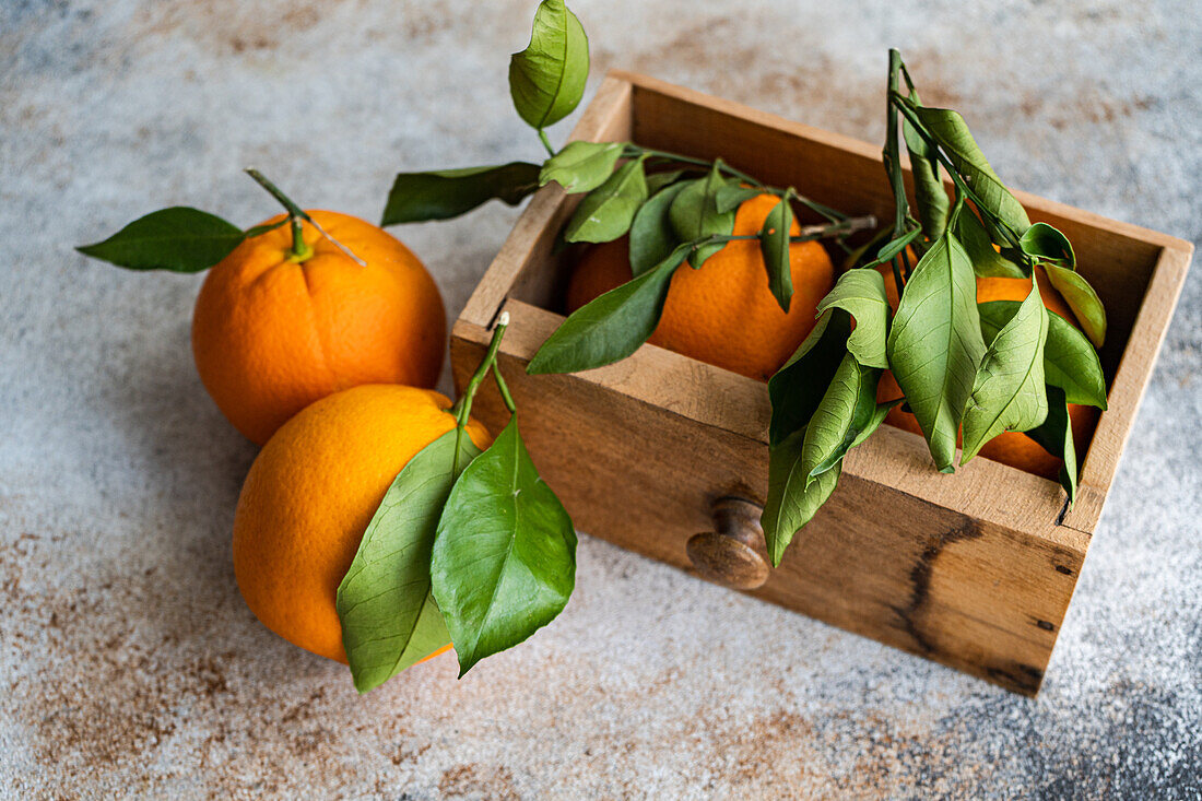 Top view of ripe oranges with fresh green leaves attached are displayed, some resting on a textured surface and others nestled in a wooden crate, evoking a sense of fresh harvest