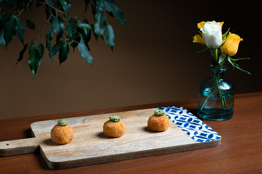 Drei goldene Arancini-Bällchen aus Reis und Käse, garniert mit Kräutern, präsentiert auf einem rustikalen Holzbrett, mit einer Vase mit gelben Rosen im Hintergrund.