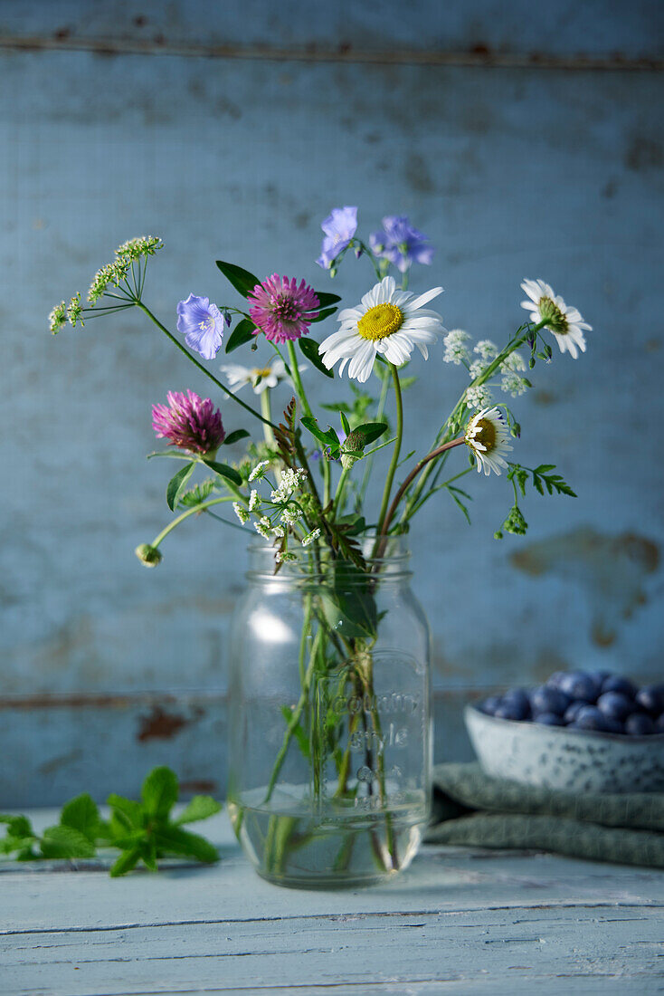 Wildblumen im Einweckglas vor blauem Holzhintergrund, Blaubeeren im Hintergrund