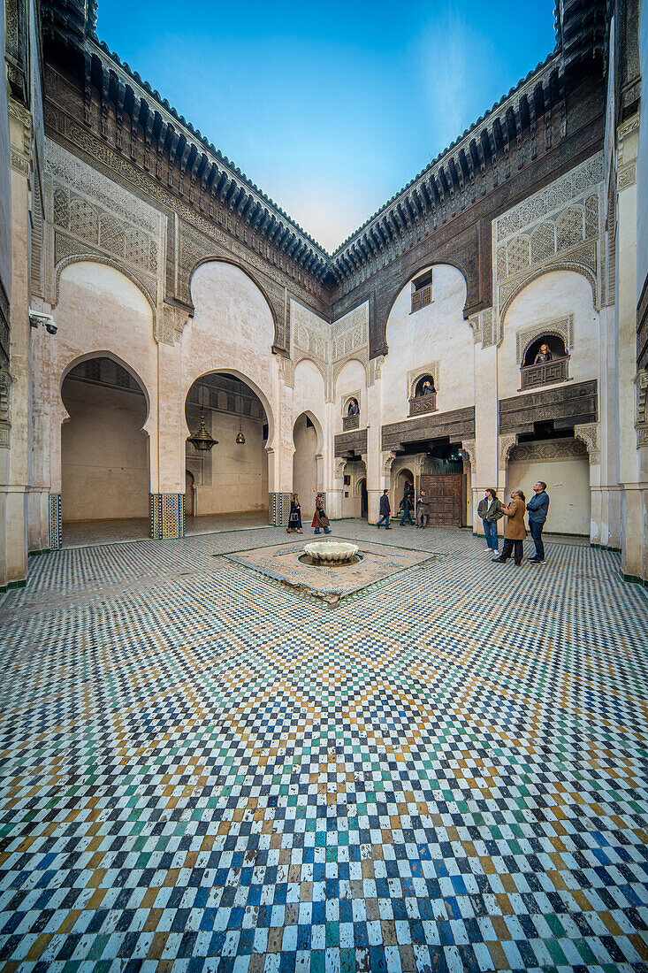 Besucher im verschnörkelten Innenhof der Madrasa Cherratine unter strahlend blauem Himmel.