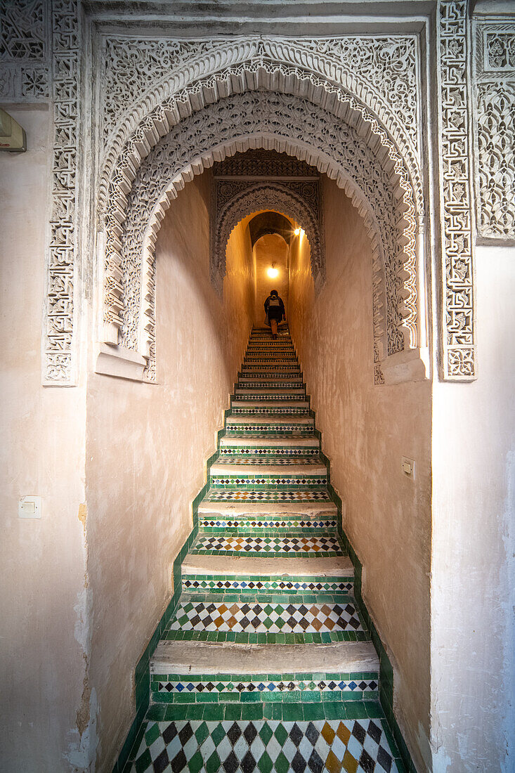 A person ascends the ornate staircase adorned with mosaic tiles in Fez's historical Cherratine Madrasa.