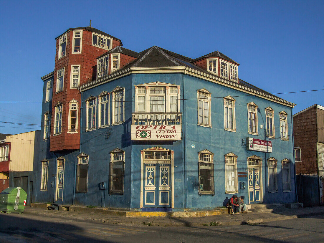 Zwei Männer sitzen vor einem bemalten, mit Metall ummantelten viktorianischen Gebäude in Castro auf der Insel Chiloe, Chile.