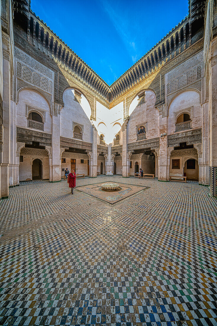 Eine Person schlendert unter strahlend blauem Himmel durch den verzierten Innenhof der Madrasa Cherratine.