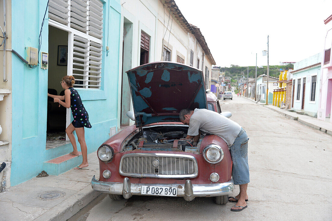 Kuba,Gibara,eine Frau betritt ihr Haus, während ein Mann den Motor seines alten amerikanischen Autos aus den 50er Jahren repariert