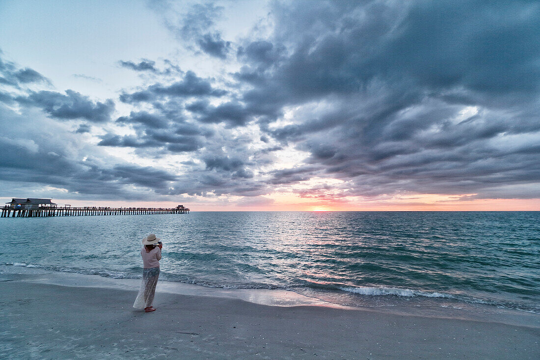 USA. Florida. Naples. Der Pier. Der Strand. Sonnenuntergang auf dem berühmten Pier. Junge Frau beim Fotografieren der Szene.