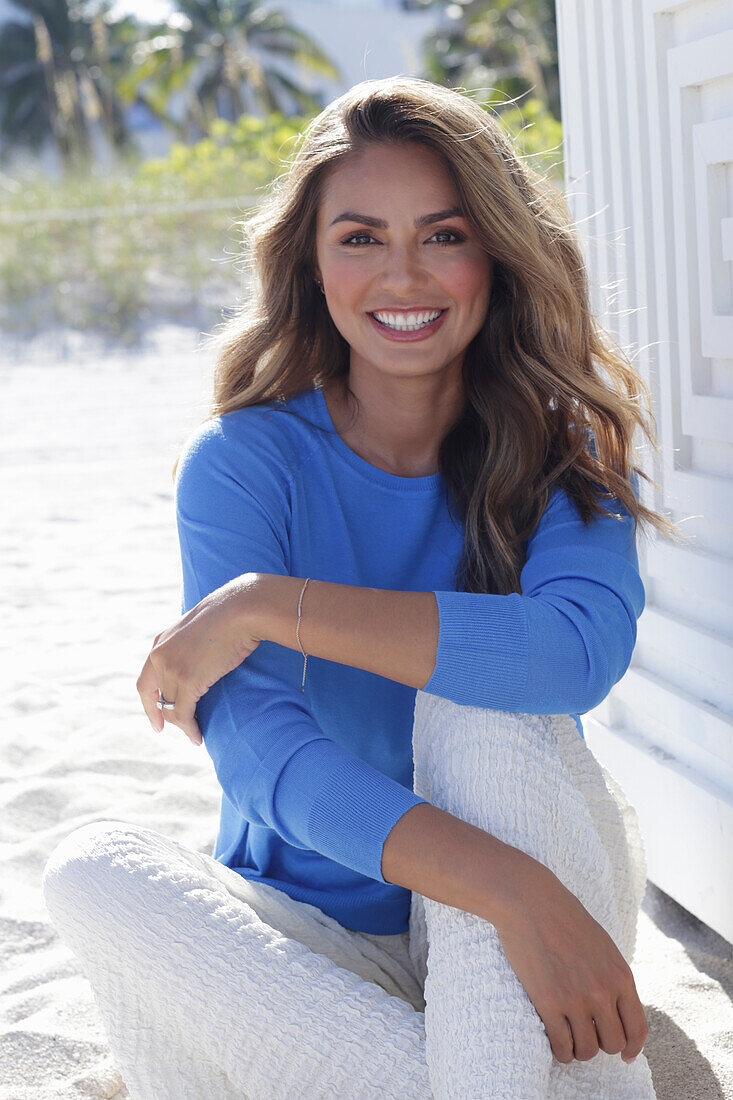 Frau in blauem Pullover und weißer Hose am Strand sitzend