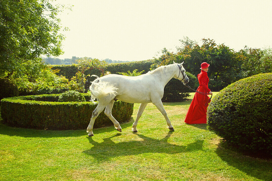 Frau in roter Reiterkleidung geht mit Pferd im Garten spazieren