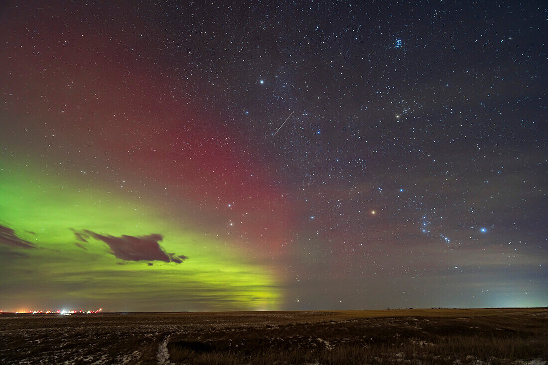 A lone meteor from the 2023 Geminid meteor shower streaks away from the radiant point in Gemini below centre amid the red aurora, with the meteor in Auriga and above Orion rising at lower right. An aurora brightens the northern sky at left, with a broad green lower curtain and red upper curtain.