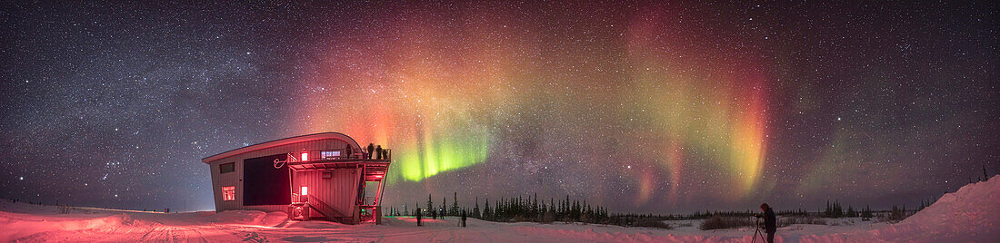 This is a panorama of a dim but colourful aurora with red curtains obvious to the camera as well as green fragments and tints of yellow, arcing across the northern sky.