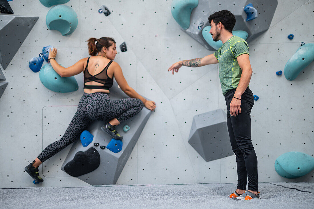 Young man teaching woman in her twenties how to climb on a climbing wall indoors