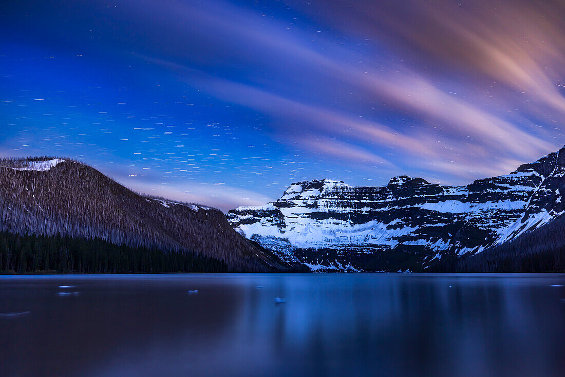 The stars and clouds trail across the sky over Cameron Lake in Waterton Lakes National Park, Alberta, and Mt. Custer across the border in Glacier National Park in Montana. The sky and mountains are illuminated by a waxing crescent Moon off frame to the right, setting in the northwest. The stars of Scorpius are rising at left. Some ice remains in this alpine lake, as this was early June 2024. Wind and the long exxposures blurs the lake surface and reflections. The hill at left is scarred by the 2017 forest fire.