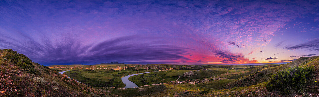 A panorama of the landscape and sky at sunset at Writing-on-Stone Provincial Park (Áísínai'pi) in Alberta, with the Milk River below winding amid the sandstone rock formations, and the Sweetgrass Hills in the distance in Montana. Note the people at far right for scale.